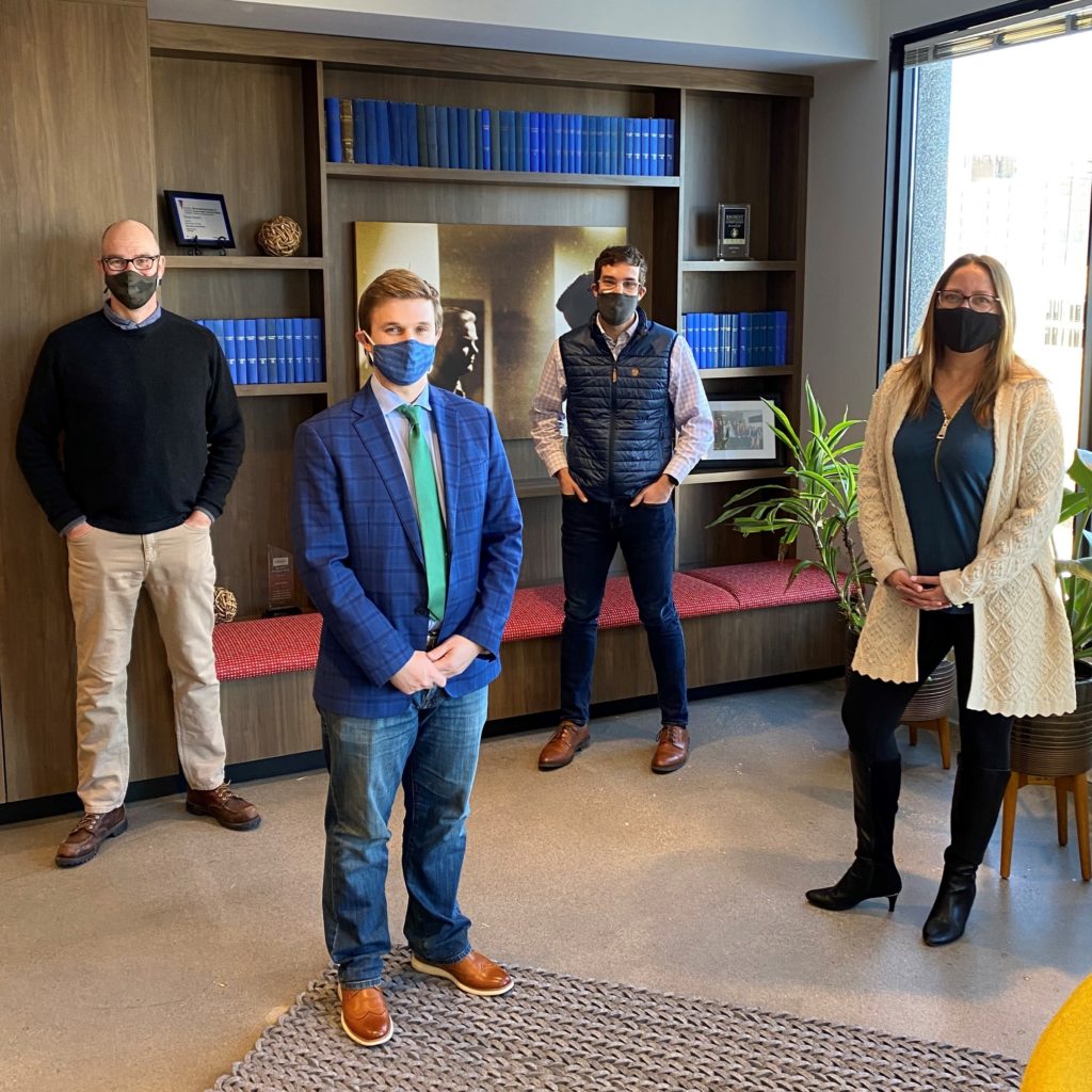 Four people wearing masks standing in an office with a bookshelf in the background