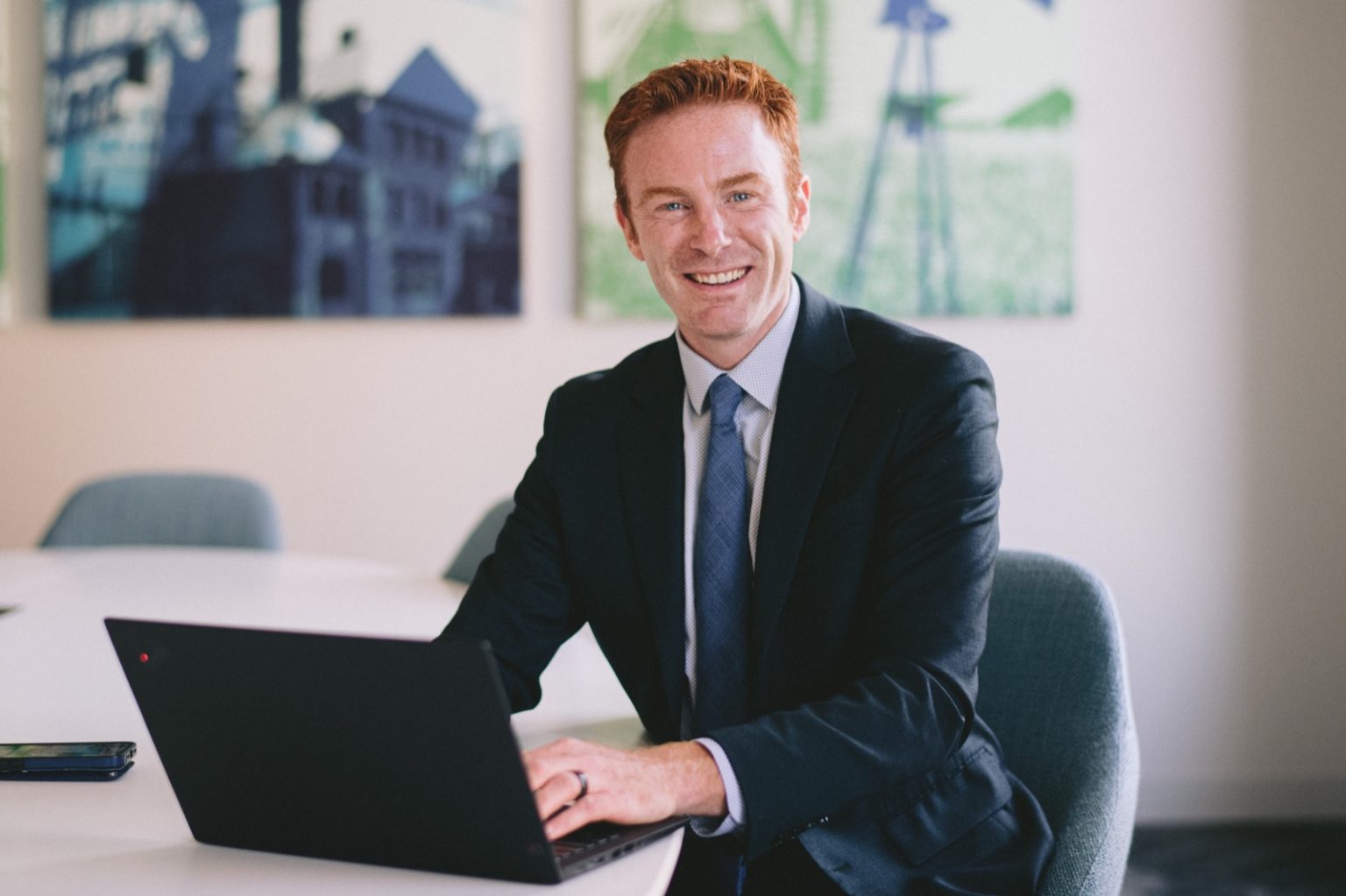 A white professional man with red hair wearing a suit and tie sits at a table with a laptop computer.