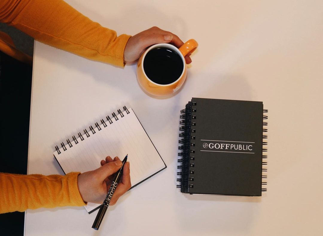 One closed notebook, one hand holding a coffee mug and one hand holding a pen about to write