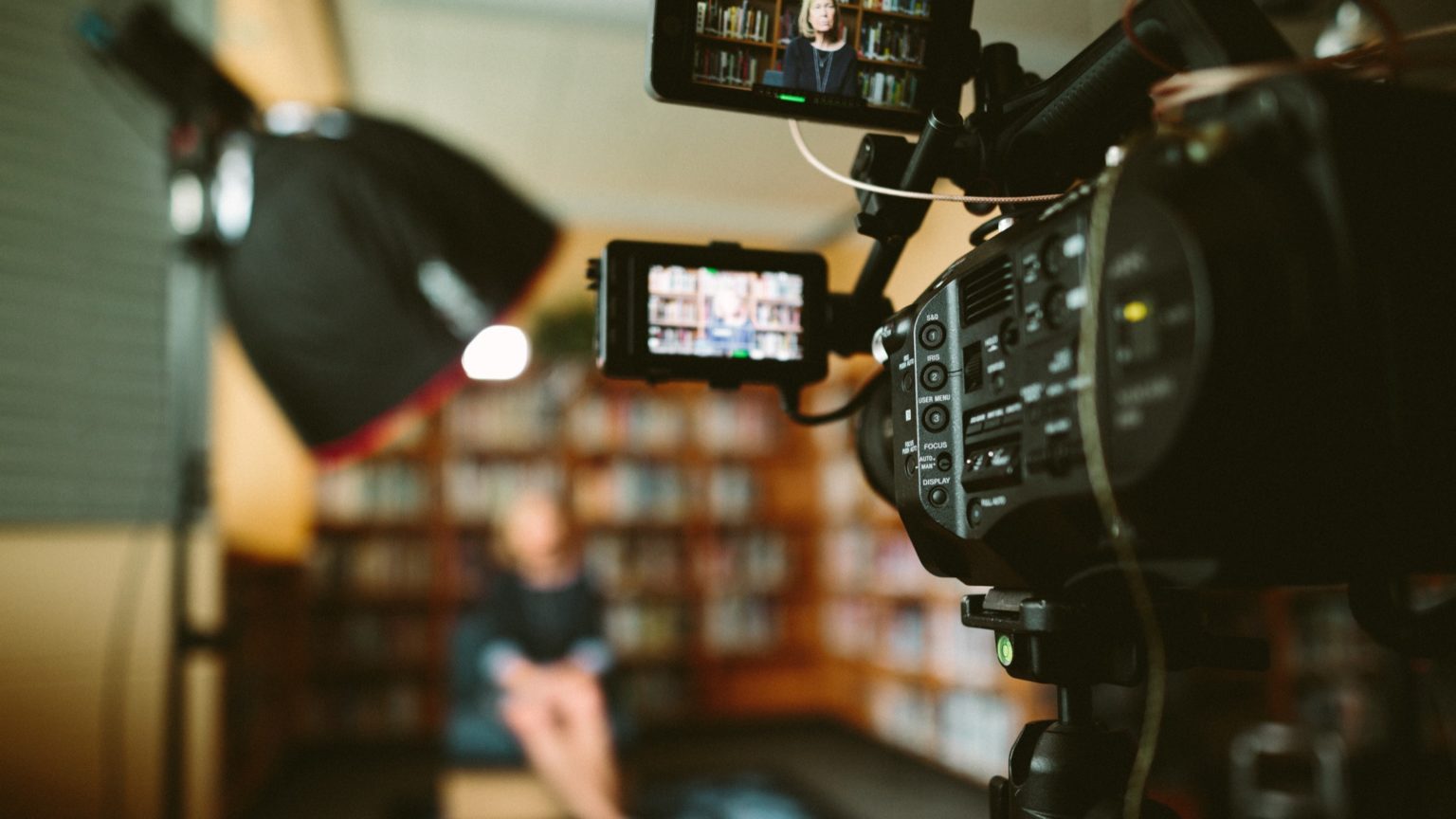 A videographer standing behind a video camera and lights recording an interview between two people