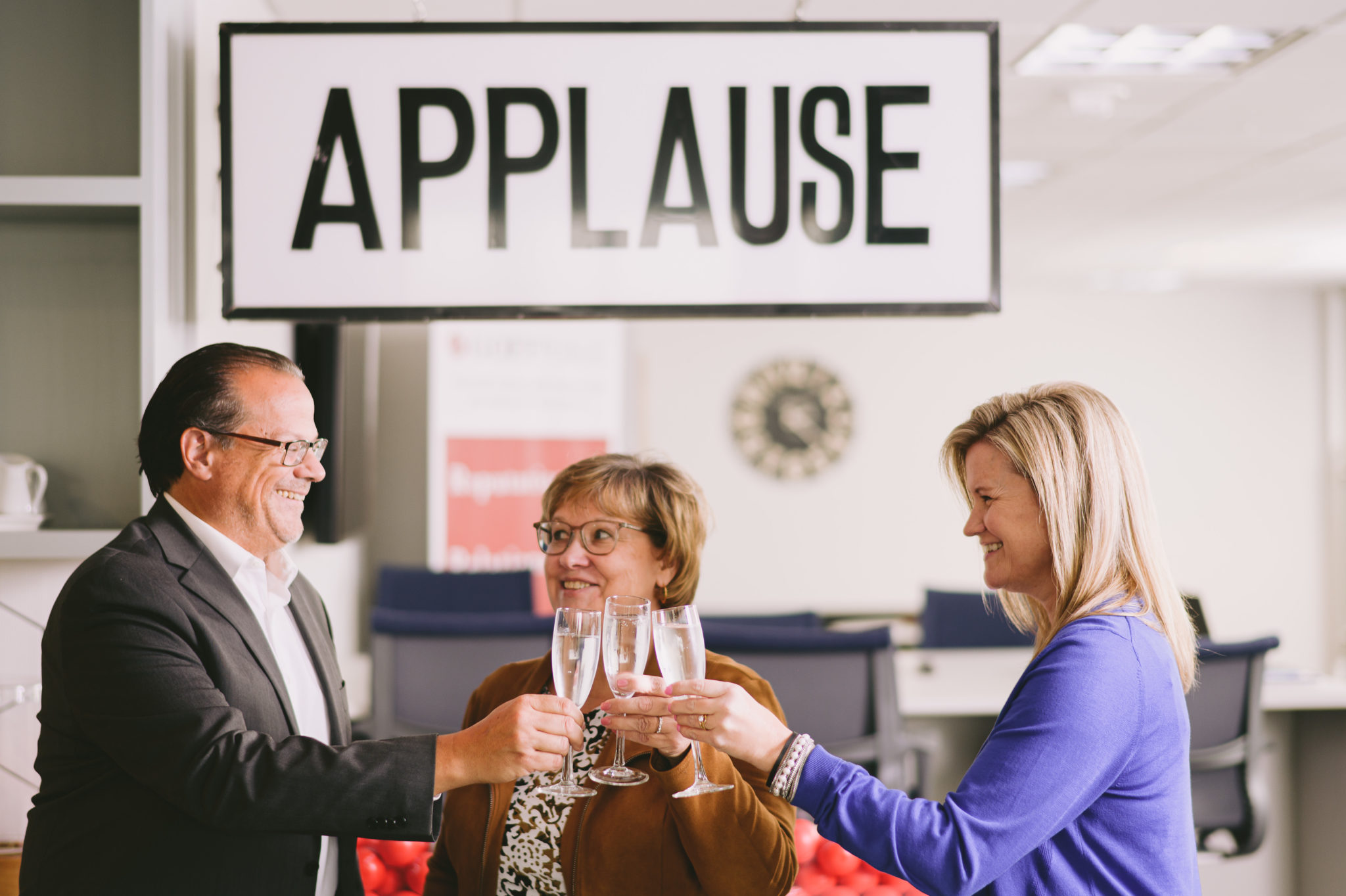 Three people standing in an office cheering glasses of champagne with a giant applause sign behind them