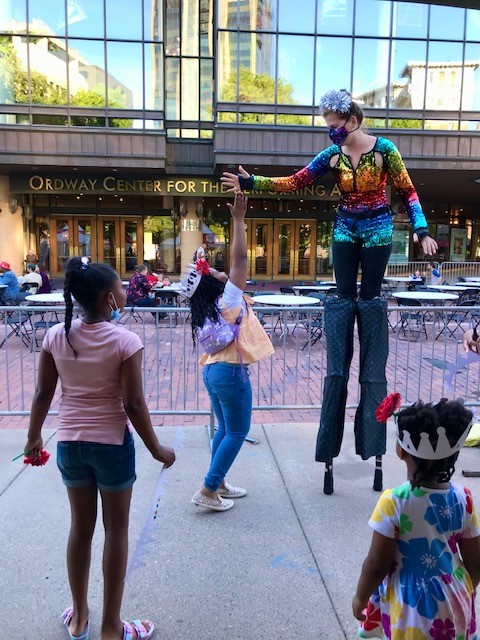 Three small children interaction with a women on stilts with the Ordway building in the background