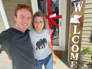 A middle-aged couple standing in front of a house with a welcome sign and bow