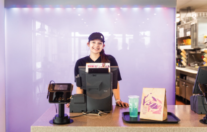 Teenager works the register of a Taco Bell with light purple background