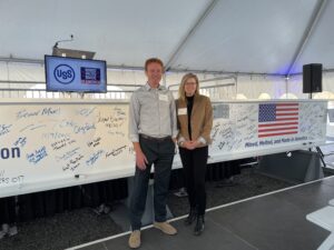 A white man and woman standing in front of a while steel construction beam with an American flag and signatures on it.