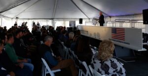 A white man speaking at a podium in front of a large seated crowds under a white tent. 