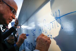 A white man with glasses holding a marker and signing a white steel construction beam.