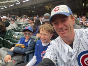 A white man sits in a stadium with his two young sons beside him. All are wearing Chicago Cubs apparel. 