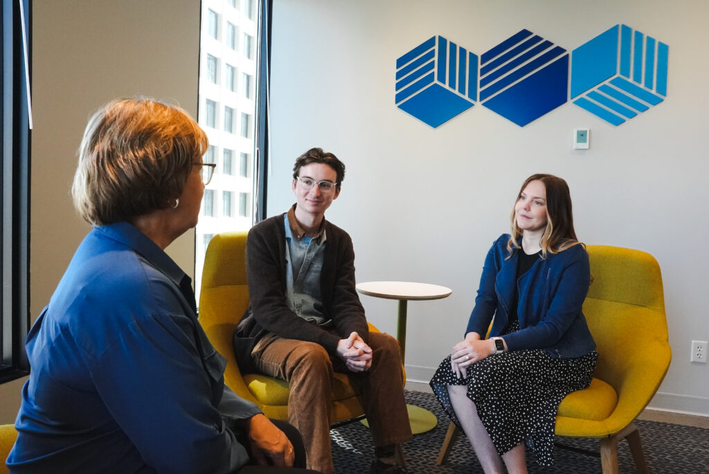 Two white professionals sit in yellow chairs, facing another person.