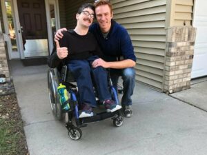 A white man in a wheel chair and a white man crouched beside him stand on the sidewalk which leads to a front door. 