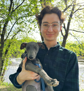 A white man with glasses holding a grey whippet puppy in a shaded outdoor scene.