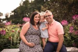 A white dad, pregnant mom and two year old girl pose for a picture in front of roses.