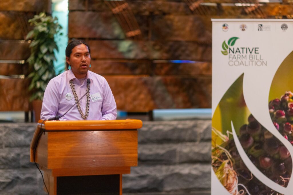 A Native man speaks at a podium next to a Native Farm Bill Coalition banner.