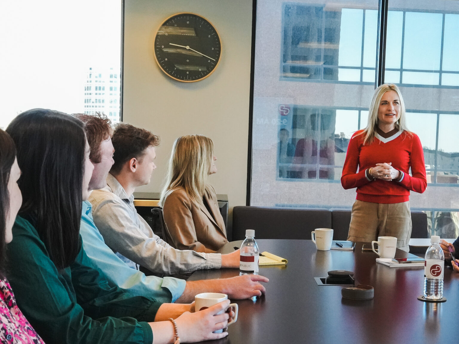 A confident blond woman speaks to a group of employees in a conference room.