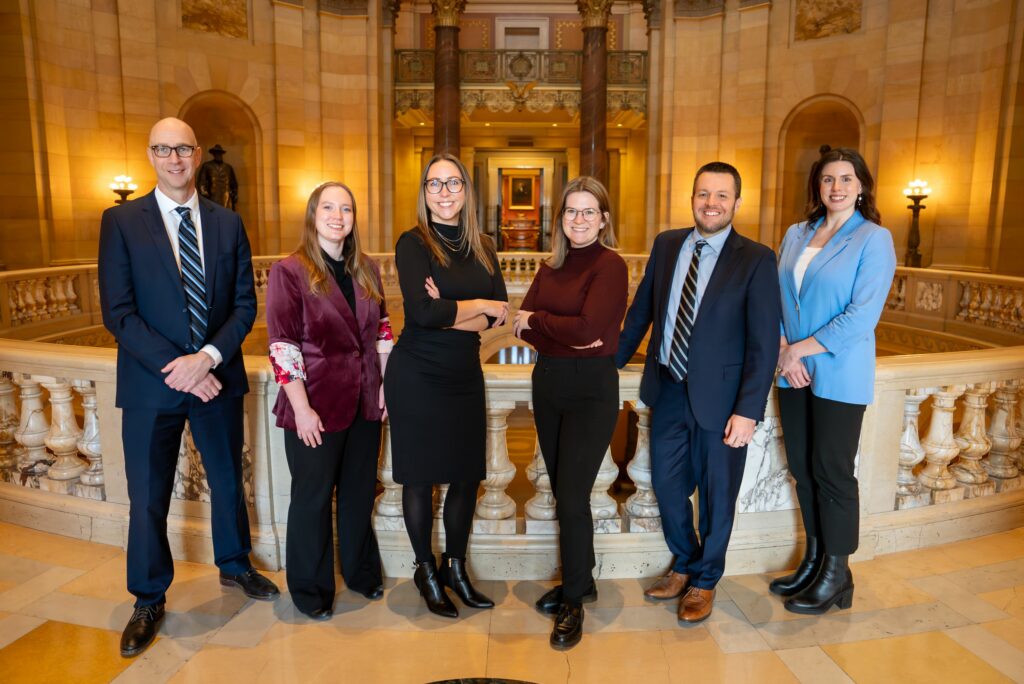 A team of people in professional clothing pose in front of the MN Capitol rotunda.