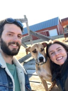 A man with dark hair and a beard takes a selfie with a dark-haired woman and a dog.
