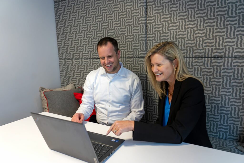 A man and woman in professional clothing smile and talk while looking at a computer screen