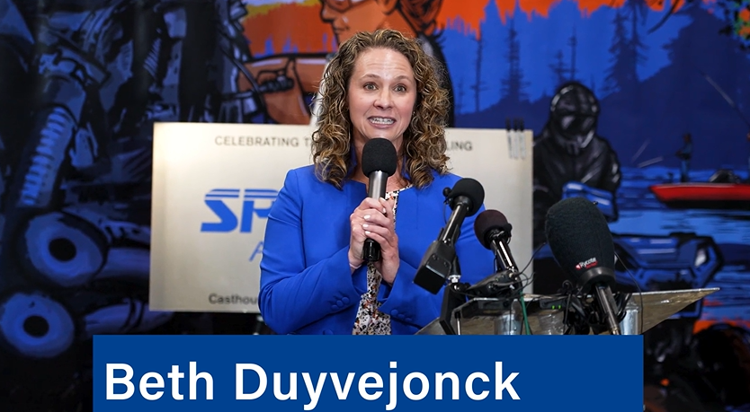 A curly-haired woman in a bright blue suit speaks into a microphone at a podium