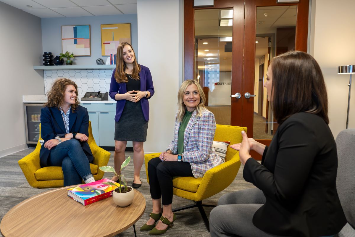 A group of women wearing professional attire talk in an office lounge area