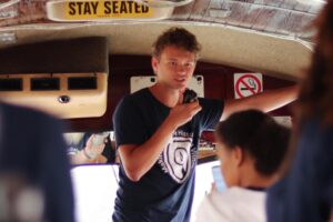 A young man speaks into a microphone at the front of a bus