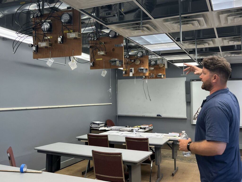 A man in a blue shirt talks while pointing at air ventilation in a classroom