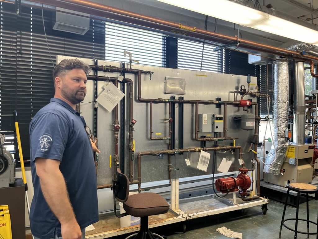 A man in a blue shirt stands in front of a pipe system