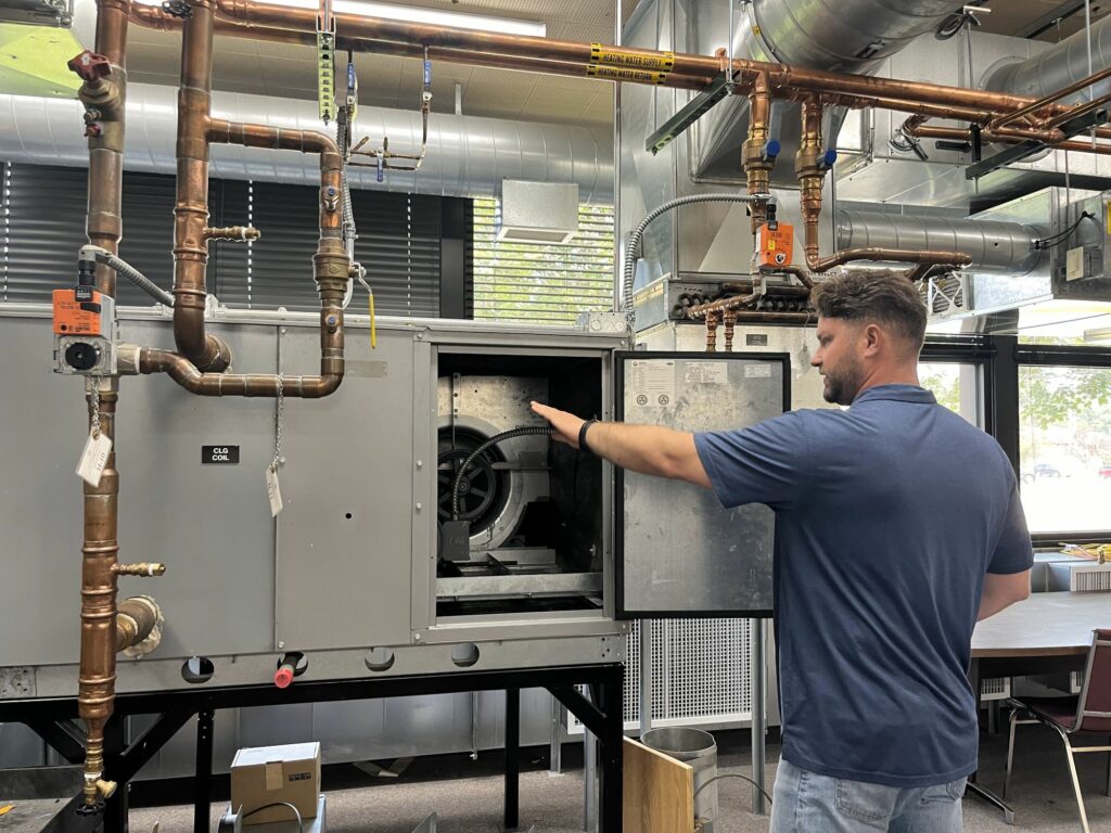 A man in a blue shirt points out parts on an air ventilation system