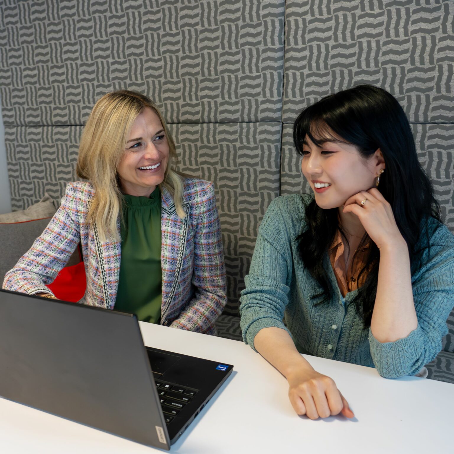 A blonde woman points to a computer screen while talking with a young Asian woman