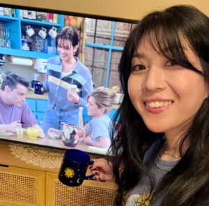 A young Asian woman poses with a TV screen playing "Friends"