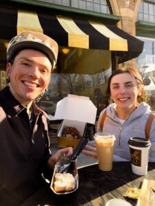 A young man wearing a bike helmet and a young woman in a sweatshirt pose with coffee and pastries outside a cafe