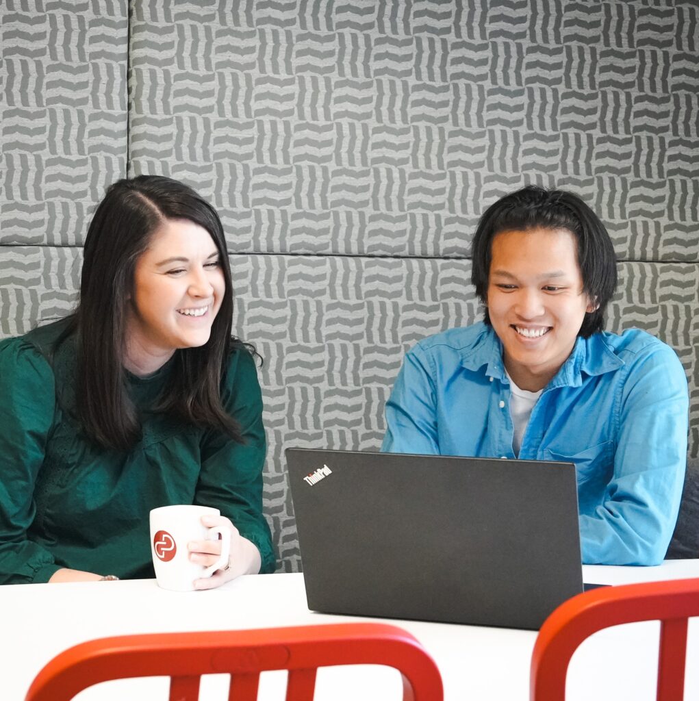 A woman with dark hair wearing a green shirt sits with a young Asian student wearing a blue button down shirt in front of a computer