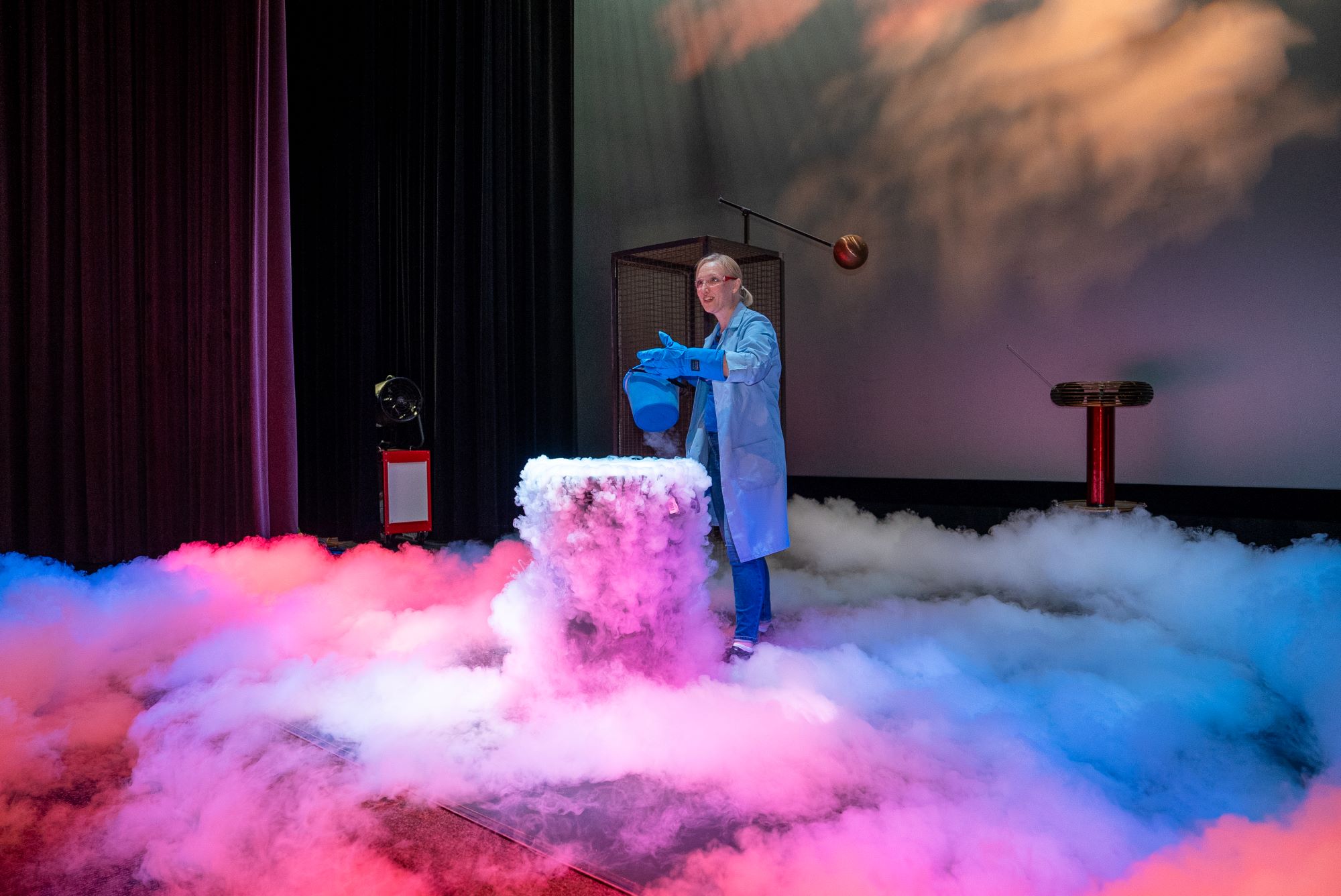 A woman scientist stands in front of a barrel on a stage filled with dry ice