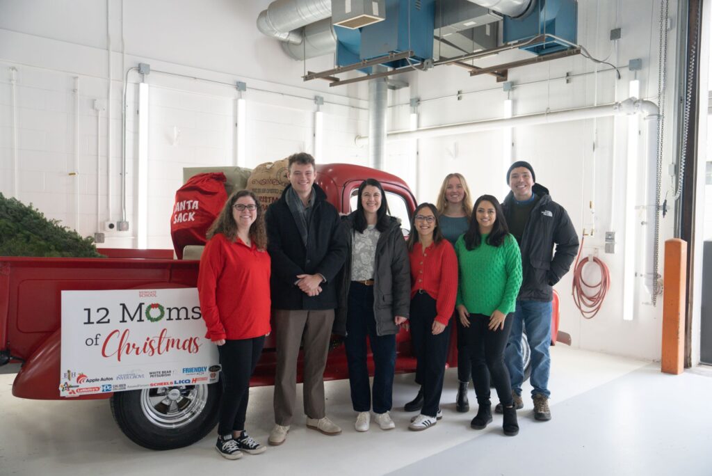 A group of people pose in front of a red pick-up truck with a sign that says "12 Moms of Christmas"