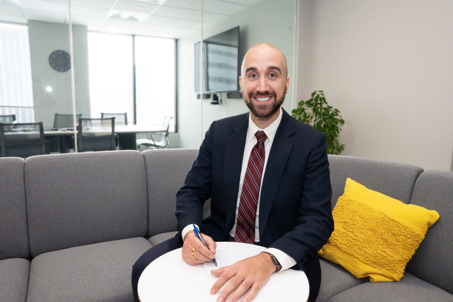 A man in a suit smiles while sitting on a couch and holding a pen and paper