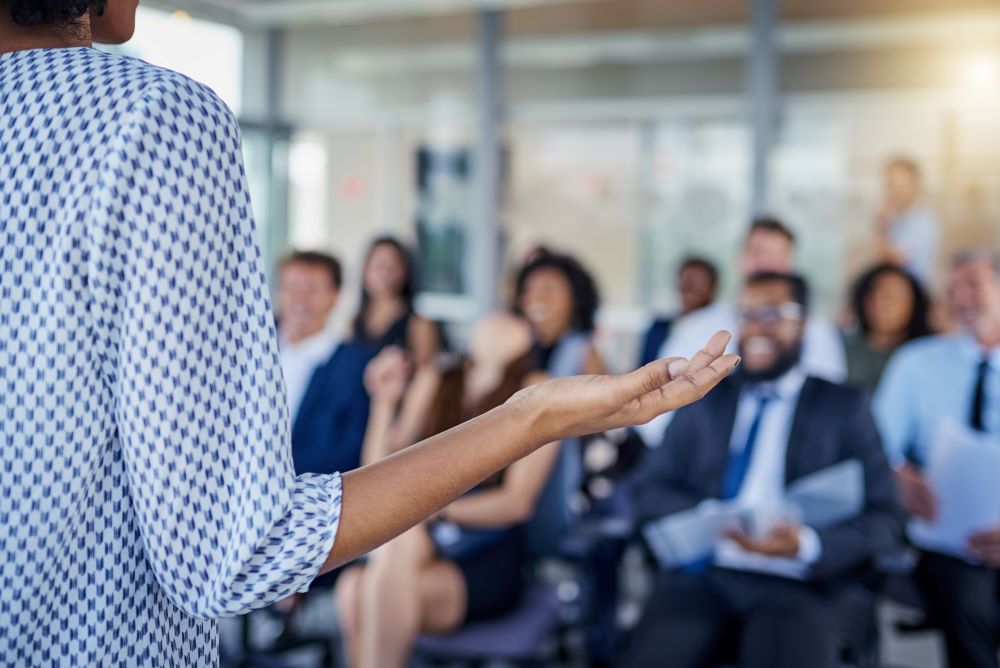 The back of a woman speaking to a group of people who are out of focus.