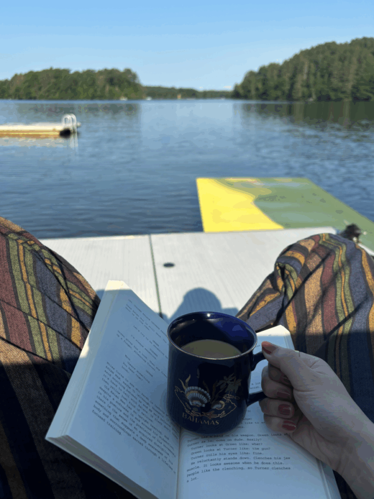 An image of a person's lap with a book and coffee mug on it overlooking a lake scene.