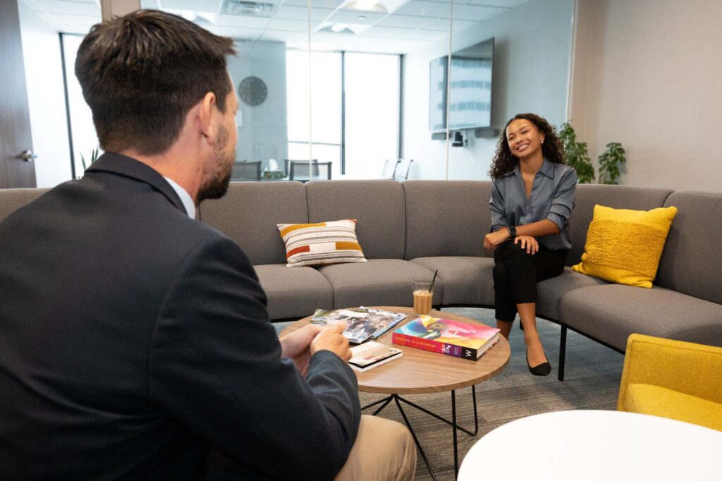 A man in a black blazer talking to a woman in a blue shirt and black pants sitting on a couch